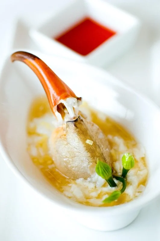 A ceramic dish holds a soup with a large shrimp on a spoon, garnished with two green onion stalks and a side of red dipping sauce.