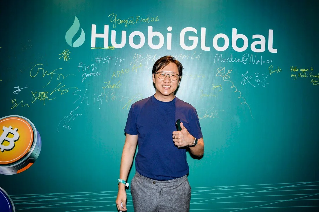 Person standing in front of a teal-colored wall with the 'Huobi Global' logo, signatures, and messages written in various colors, giving a thumbs-up gesture.