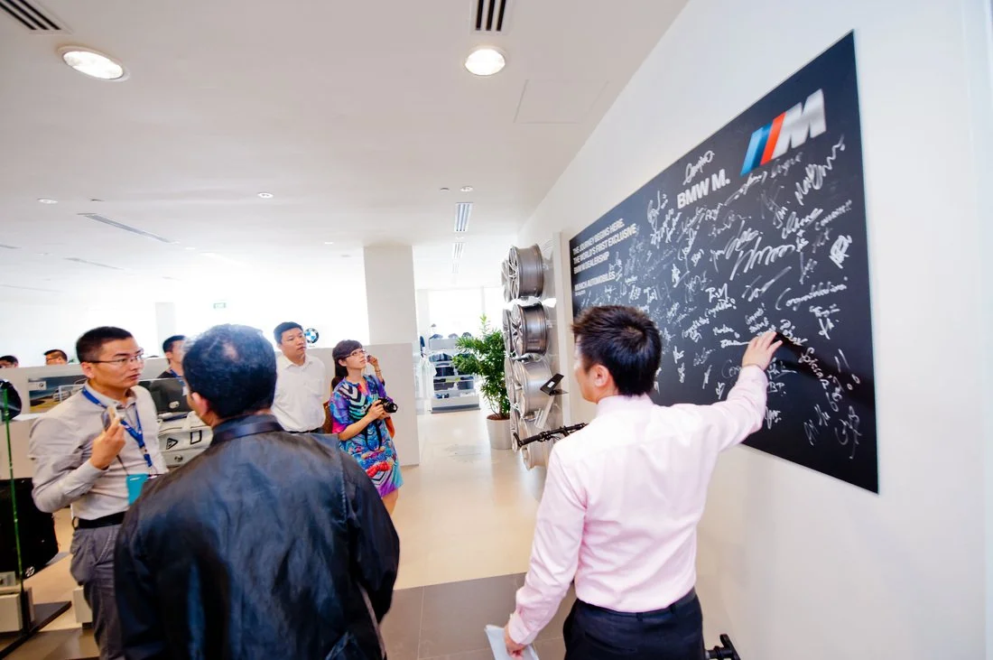Group of people in a BMW showroom, some taking photos, one writing on a blackboard with signatures, BMW logos and decorations on the wall.