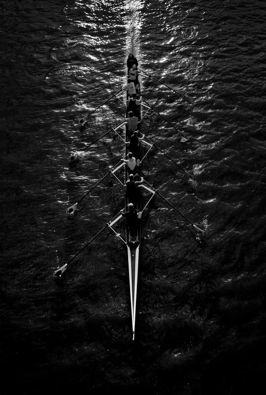 A black and white aerial view of a rowing team on a river, with the water reflecting light.