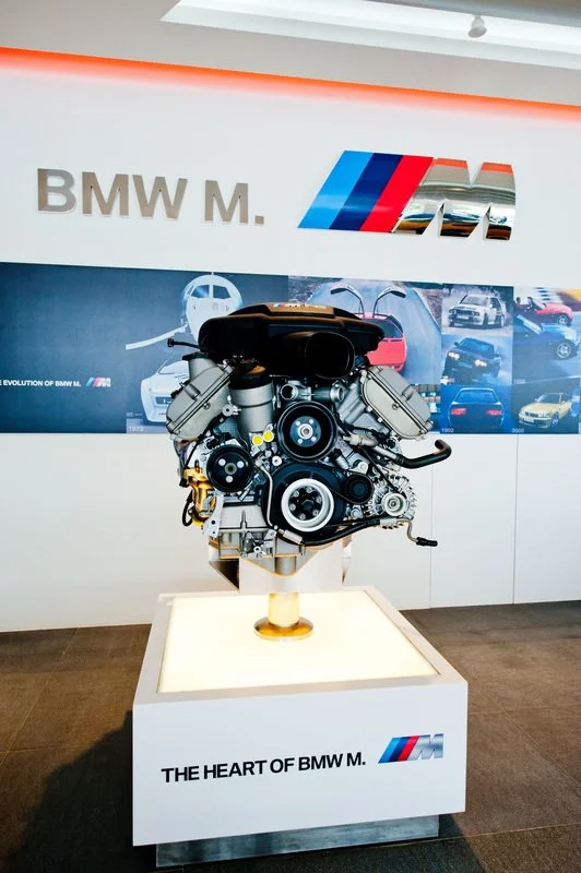 A BMW M engine displayed on a white pedestal in a showroom with the BMW M logo and a banner of various BMW cars in the background.