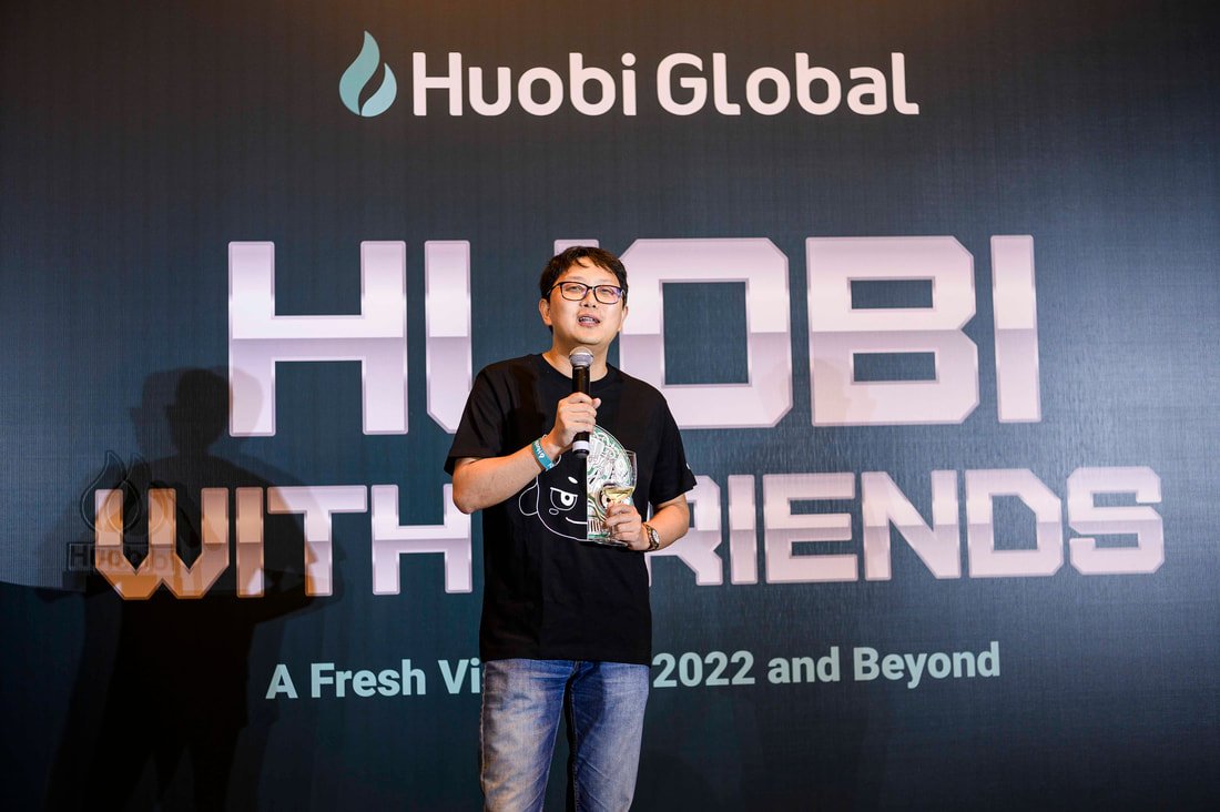A man speaking at a conference, holding a glass trophy and a microphone, standing in front of a large screen displaying 'Huobi Global' and 'HUIDI WITH FRIENDS, A Fresh View 2022 and Beyond'.