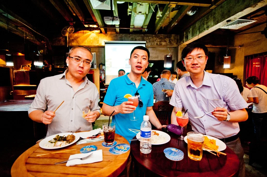 Three men standing behind a round table at a restaurant, holding drinks and food, with a lively atmosphere and other patrons in the background.