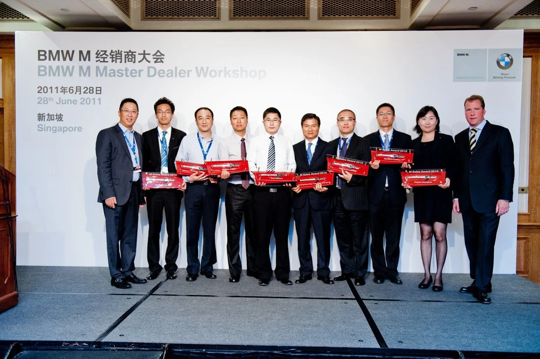 Group of nine people standing on stage at a BMW event, holding red boxes and posing for a photo.
