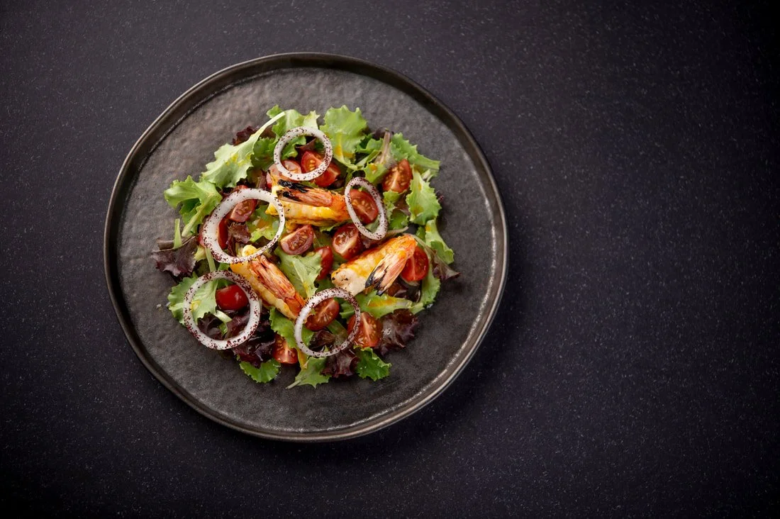 Salad with shrimp, cherry tomatoes, onion slices, and mixed greens on a black plate.