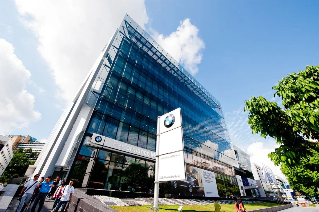 Modern glass office building with BMW logo, sign reads 'Performance Motors', and a few people walking outside.