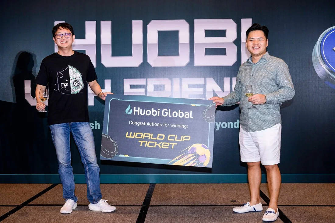 Two men smiling and holding glasses of wine stand on either side of a large sign that reads "Huobi Global" and "Congratulations for winning WORLD CUP TICKET." The background has the words "HUOBI WORLD FRIENDS."