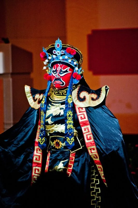 Traditional Chinese opera performer in elaborate costume and painted face mask with red, blue, white, and black designs.