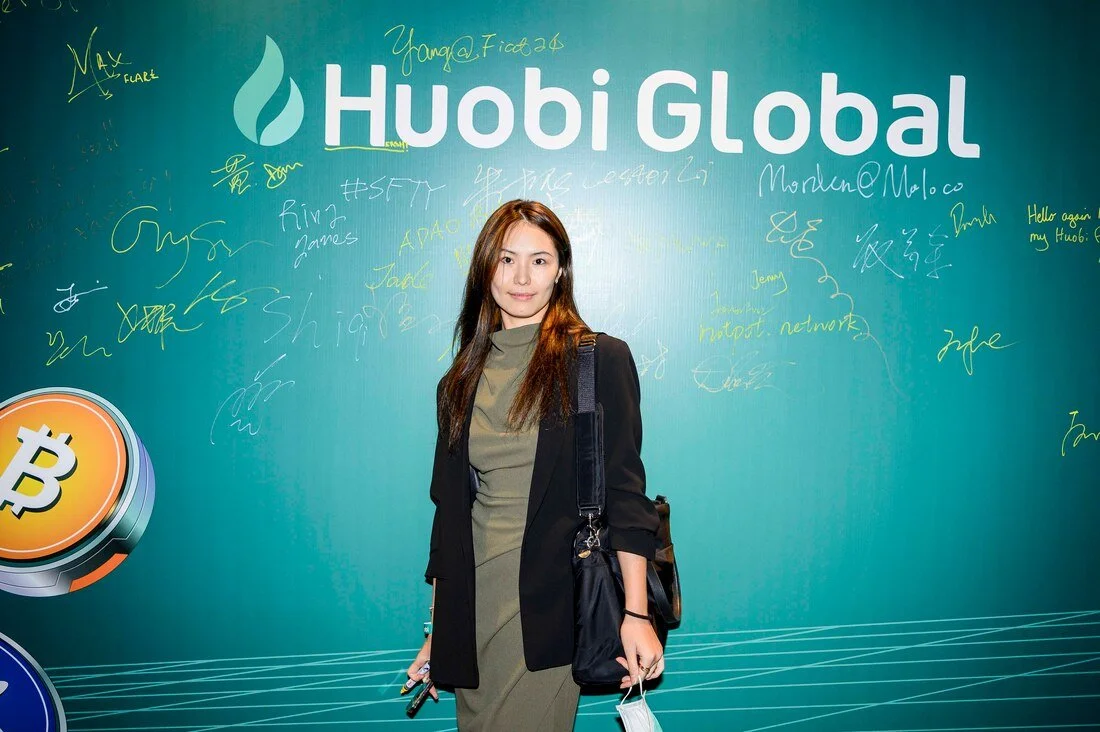 Young woman standing in front of a blue Huobi Global promotional backdrop with signatures and writings, holding a phone and face mask.