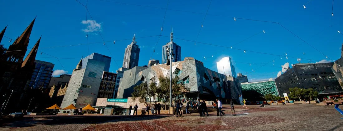 A city square with modern buildings, a clear blue sky, and string lights overhead. People are walking and sitting on benches.