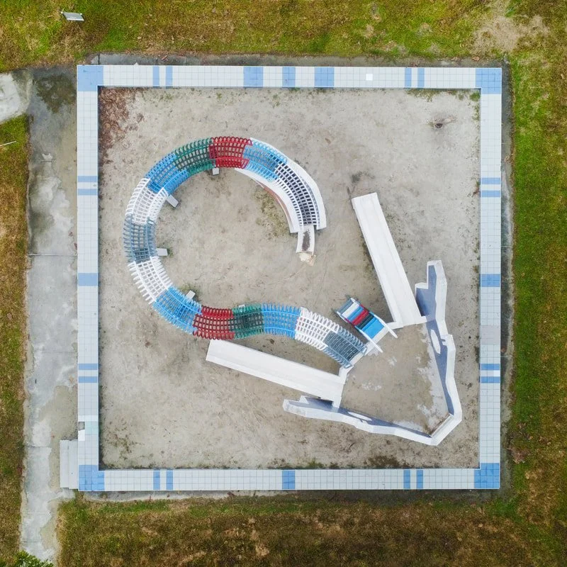 A bird's-eye view of a small, deserted water park with a winding, multi-colored water slide and shallow pool area, enclosed by a tiled border and surrounded by grass.