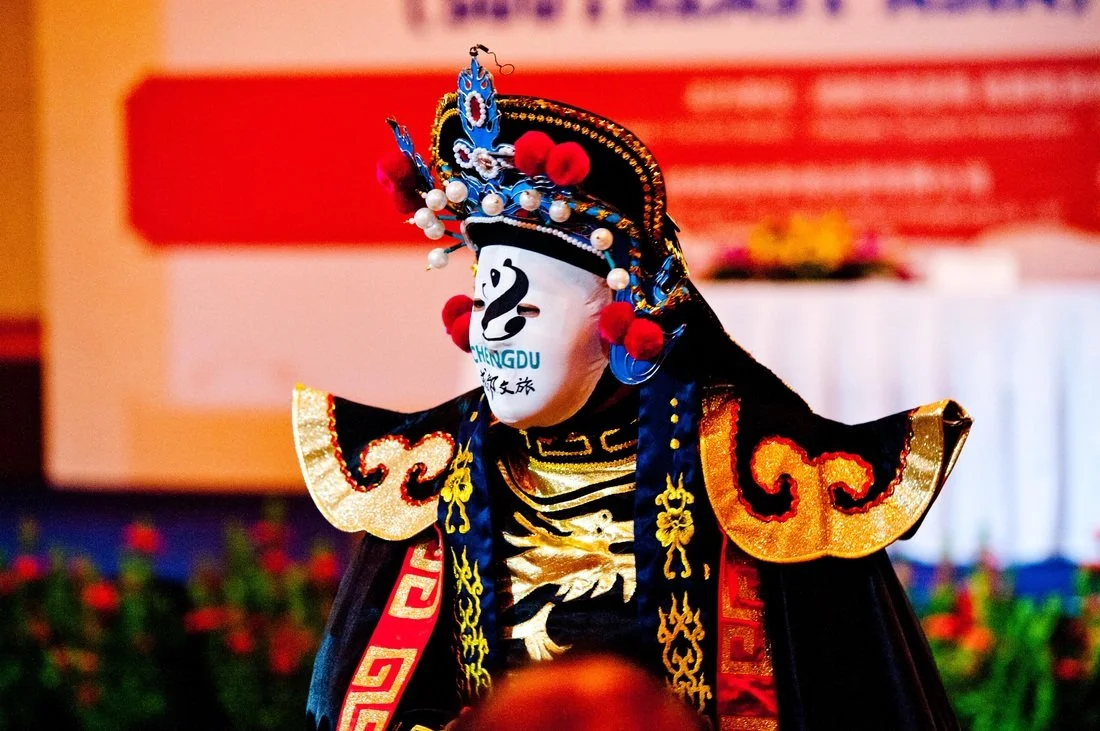 A performer in traditional Chinese opera costume with a painted face mask and elaborate headdress.