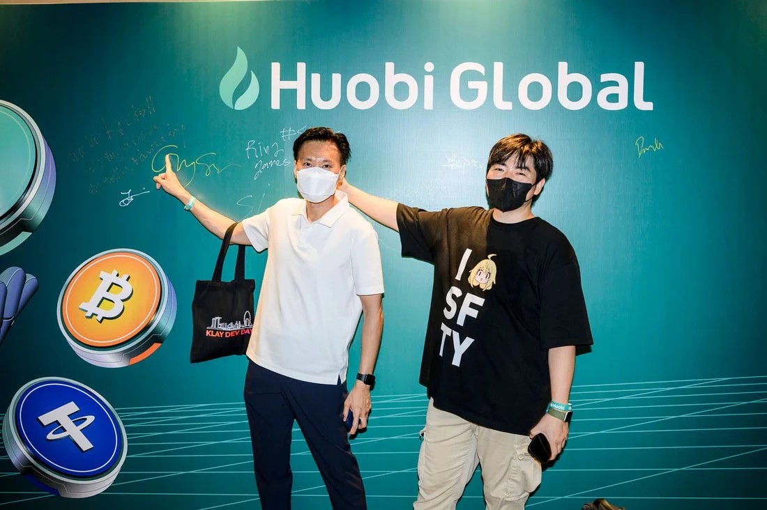 Two men wearing masks, one in a white shirt and the other in a black T-shirt with anime character, stand in front of a blue-green backdrop with the logo for Huobi Global, holding up a pointing gesture and signing on the wall.