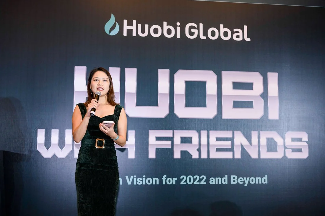 A woman in a black dress speaking into a microphone on stage, with a screen behind her displaying the Huobi Global logo and text about a conference in Dubai.