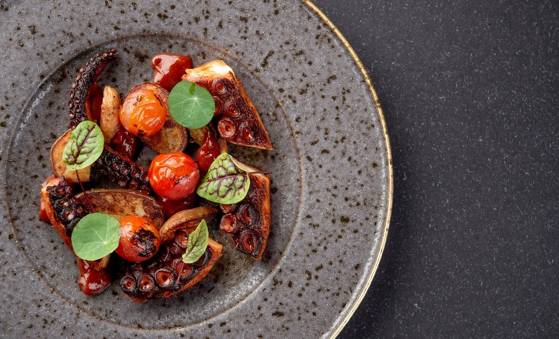 Grilled octopus tenticles garnished with cherry tomatoes and microgreens on a dark speckled plate.