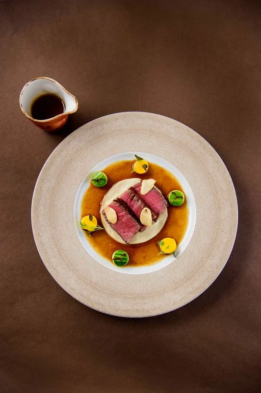 A plated gourmet dish with slices of seared beef topped with garlic and sauce, garnished with colorful vegetables, and served with a small pitcher of sauce on the side.