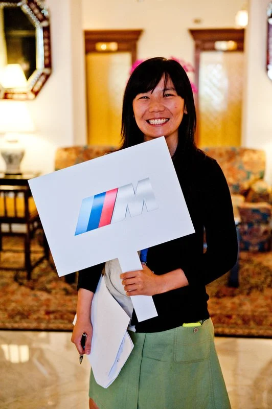 A smiling woman holding a sign with the BMW M logo in a cozy, well-lit room.
