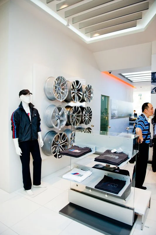 Display of car rims on a white wall, a mannequin dressed in a jacket and pants with a face mask, and two customers in striped shirts browsing in a showroom.