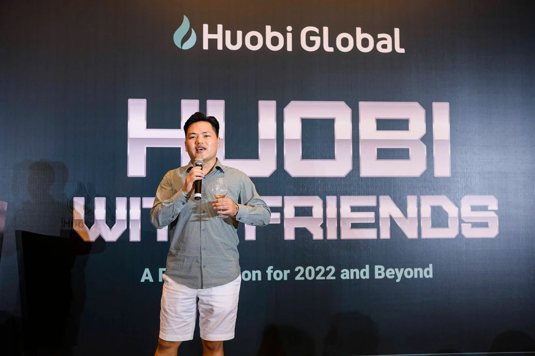 A man holding a glass and speaking into a microphone on stage at a Huobi Global event with a large screen behind him displaying the logo and text.