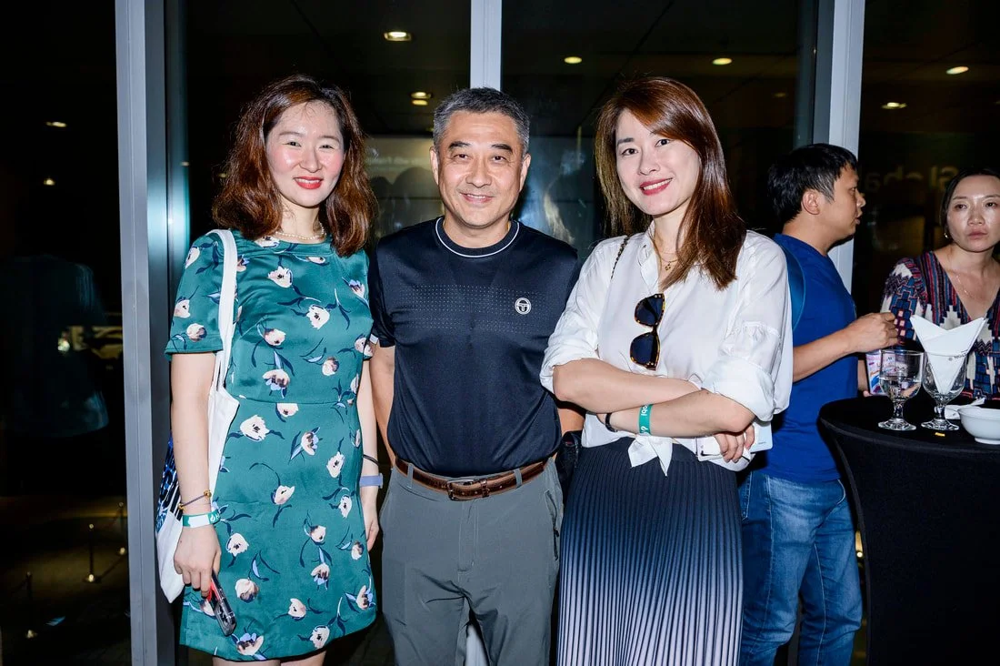 Three people pose for a photo at a social event, standing in front of large glass windows with a nighttime city view.