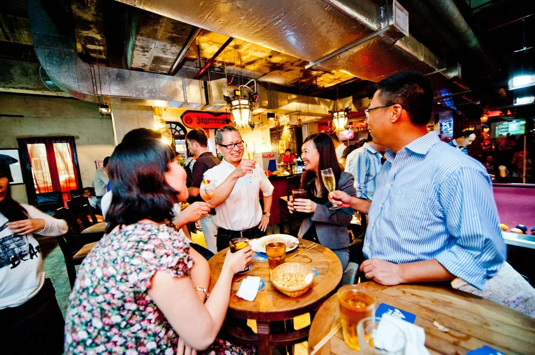 Group of friends enjoying drinks and chatting at a bar or pub, with warm lighting and a lively atmosphere.