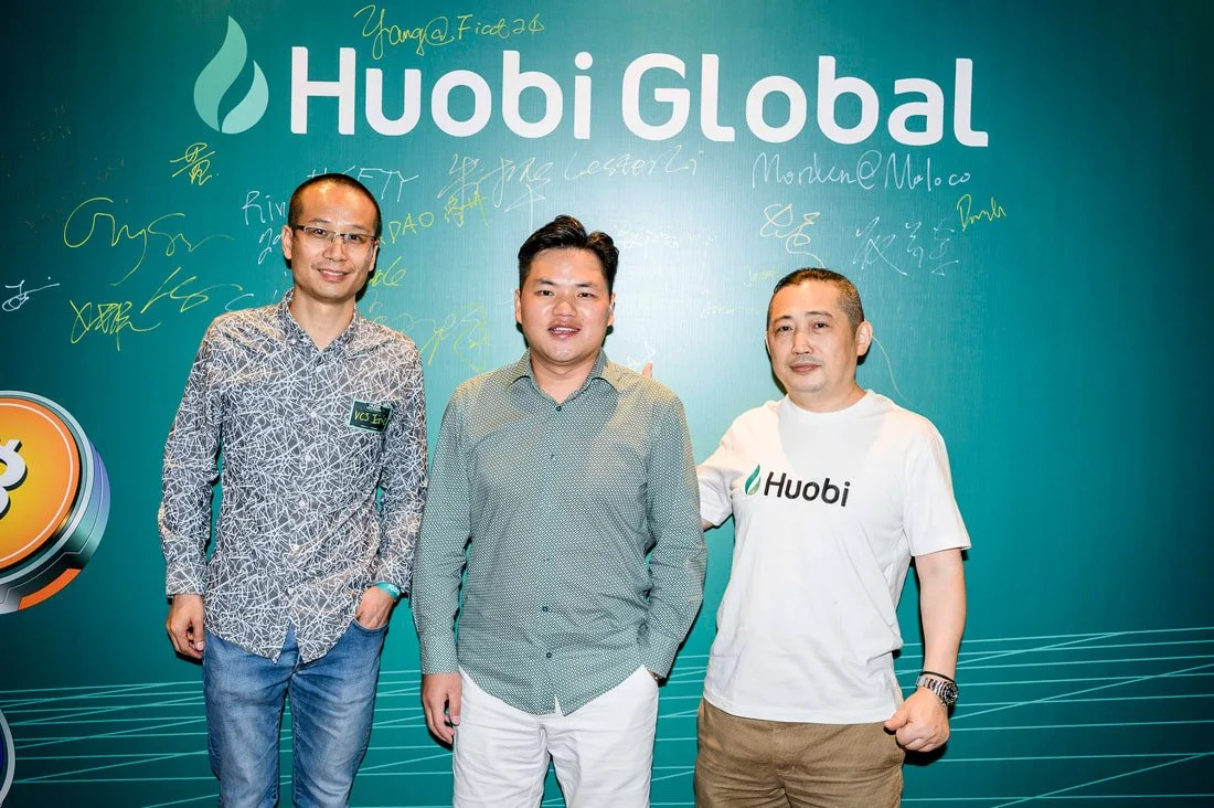 Three men standing in front of a teal Huobi Global backdrop with signatures and handwritten notes, smiling at the camera, one wearing a Huobi T-shirt.