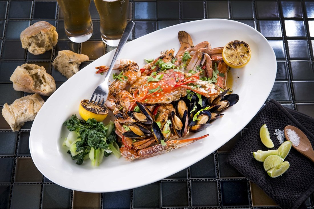 Seafood platter with crab, mussels, and shrimp on a white plate, garnished with lemon and greens, accompanied by bread, lime wedges, salt, and two glasses of beer.