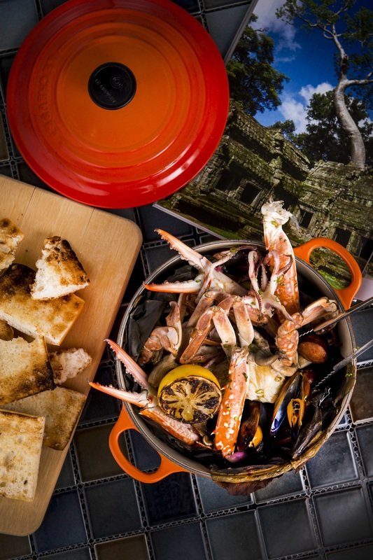 A pot of fresh seafood including crab legs, shellfish, and shrimp, with a lemon slice inside, set on a black patterned table. Nearby, there is a wooden cutting board with pieces of toasted bread, and a red bowl with a lid is placed above the seafood.