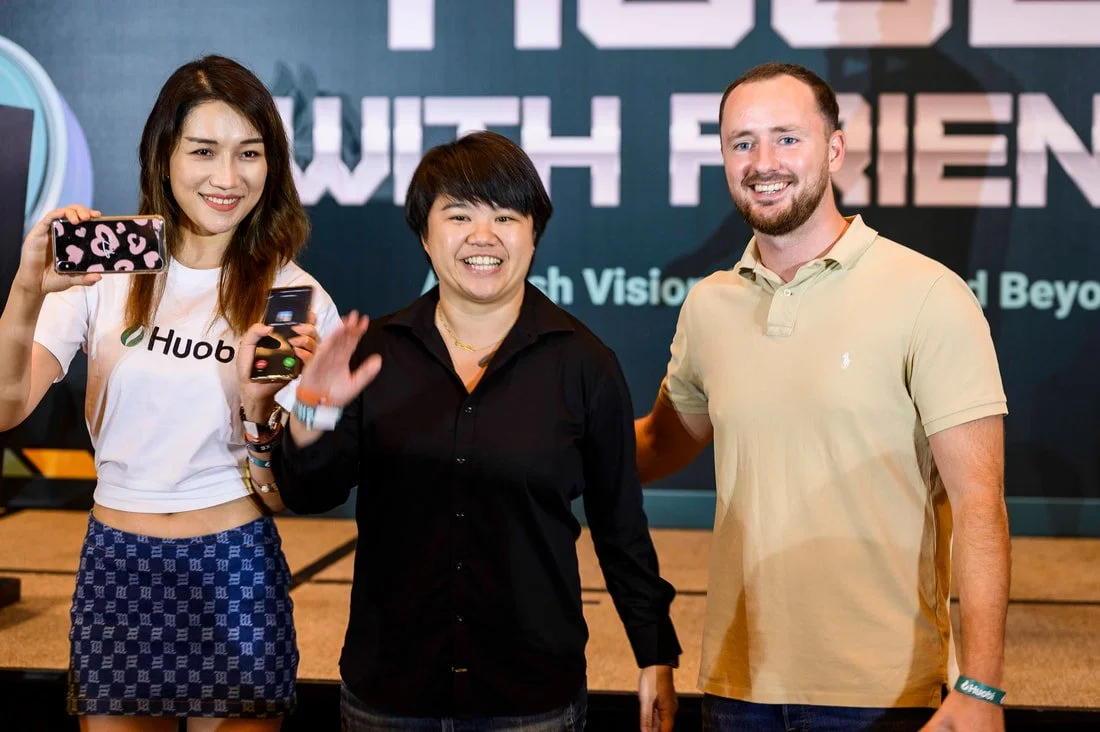 Three people standing together at a conference, smiling for the photo. The woman on the left is holding a smartphone, the woman in the middle is waving, and the man on the right has his arm around her. The background displays a large screen with part