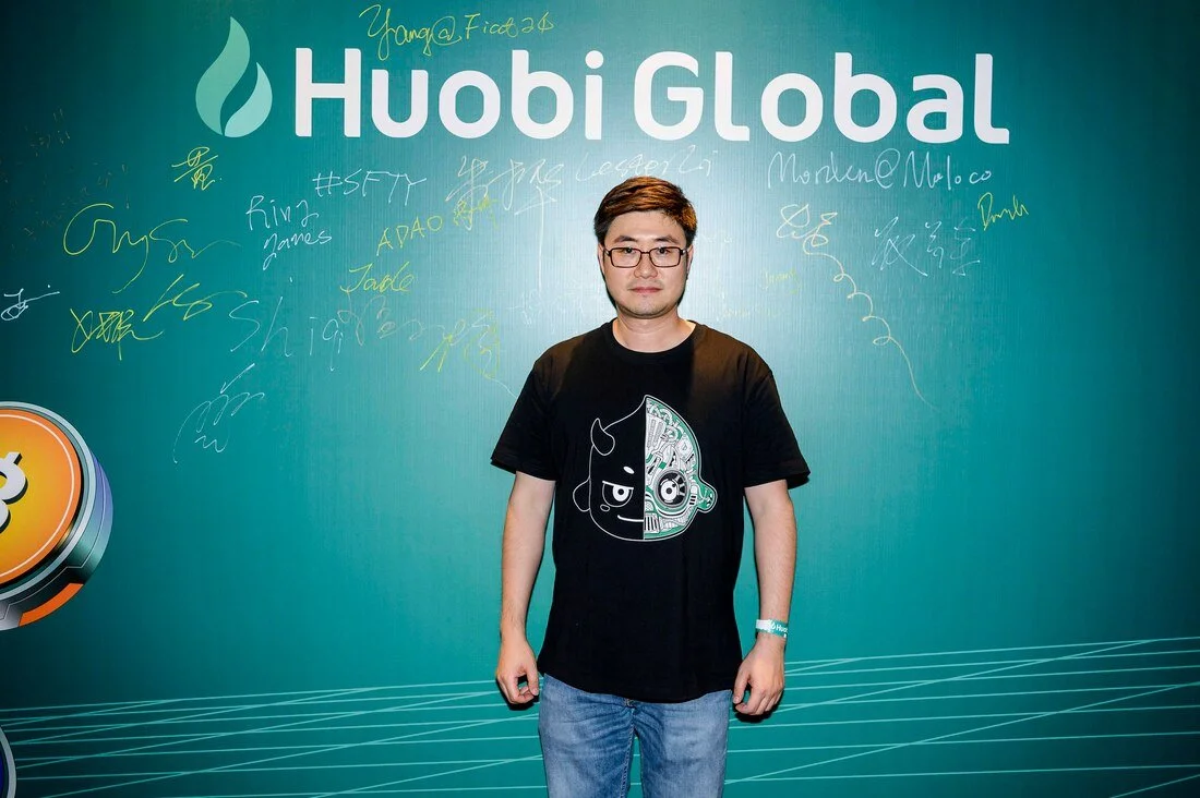 A man in glasses wearing a black T-shirt with a half cartoon face design standing in front of a teal Huobi Global backdrop with signatures and scribbles.
