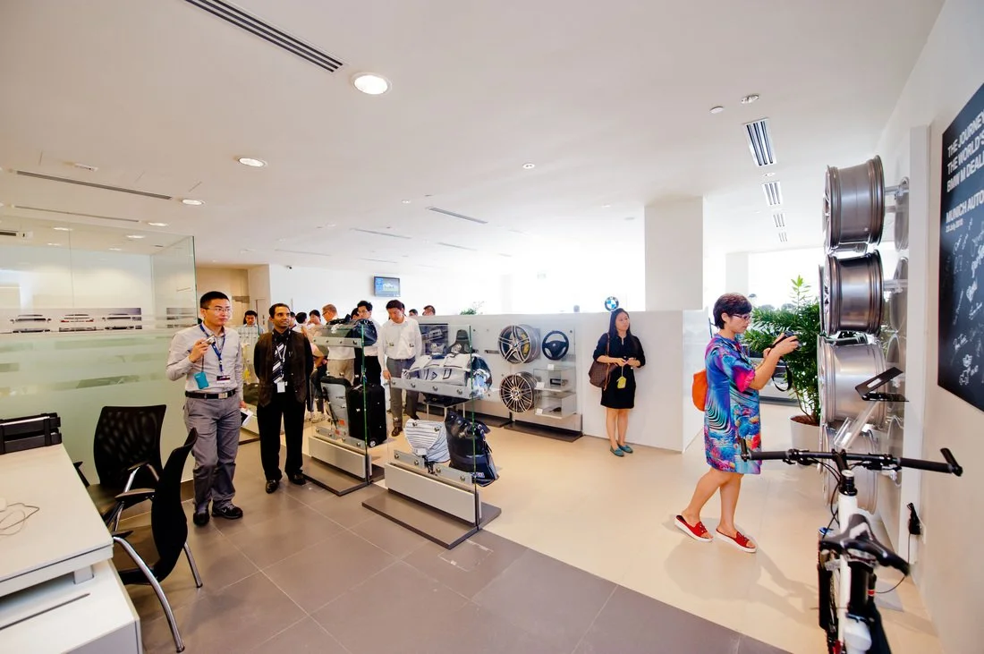 People browsing automotive parts and accessories in a showroom or retail store, with displays of wheels, steering wheels, and related items on white shelves.