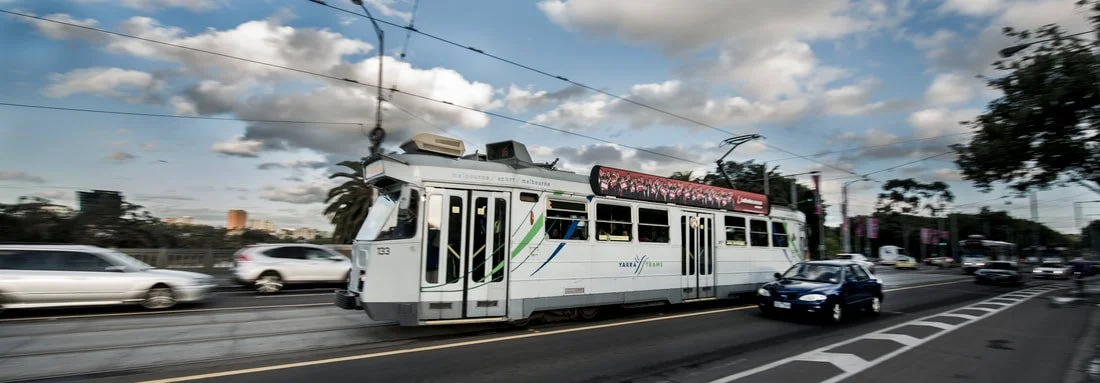 A light rail train moving along tracks on a city street with cars and traffic, under a partly cloudy sky.