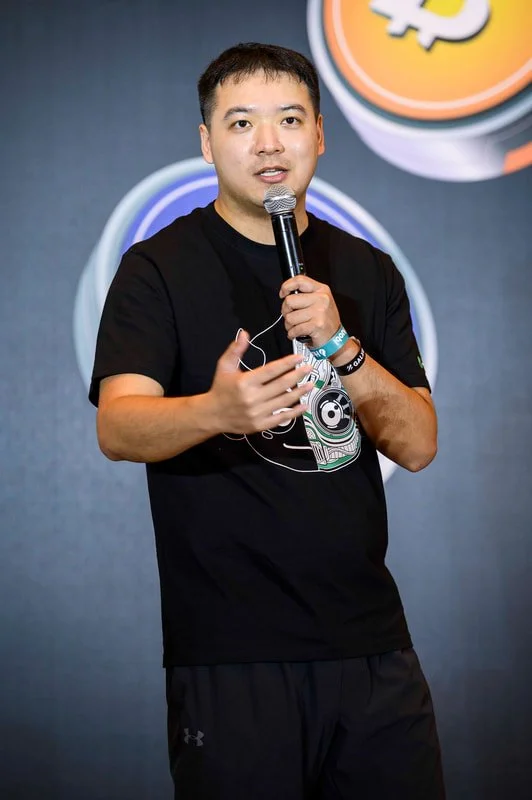 A man wearing a black T-shirt with a graphic design and black pants, holding a microphone and speaking at an event, with a background featuring colorful circles and cryptocurrency logos.