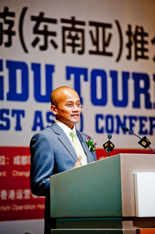 A man in a suit and glasses speaking at a podium with a microphone in front of a large conference banner. The banner has Chinese characters and English text, mentioning 'Education Tour' and 'Conference'.