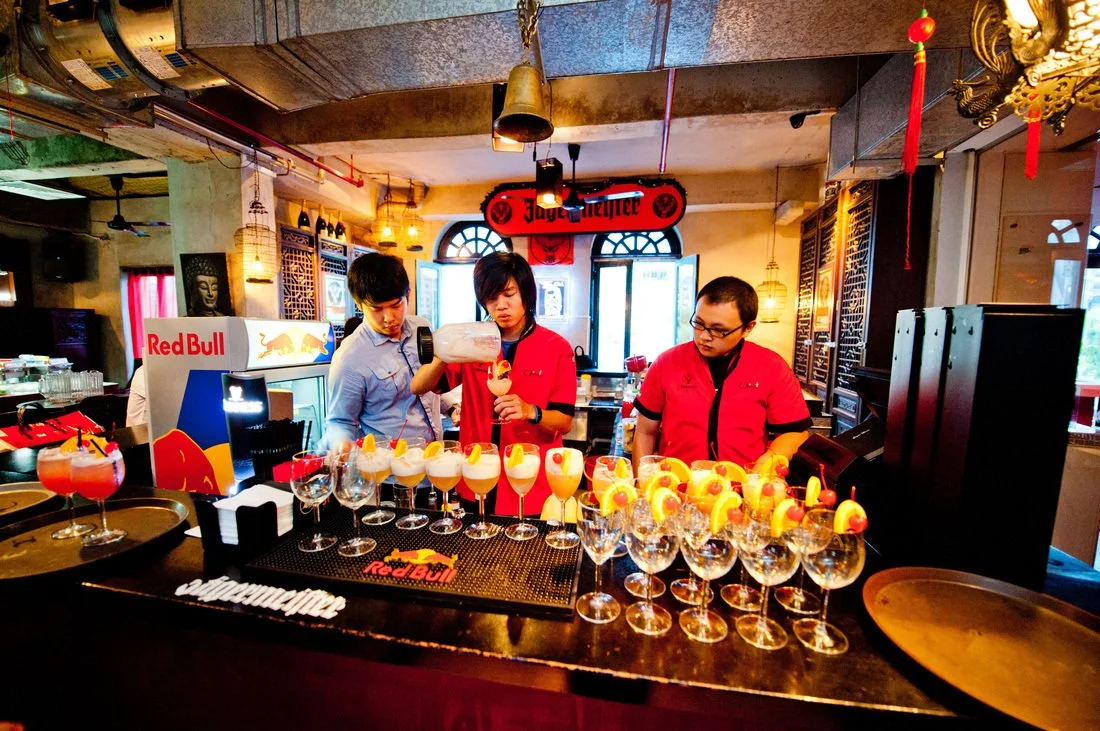 Barista preparing colorful cocktails at a bar with Red Bull branding, surrounded by glasses with garnished beverages, in a dimly lit indoor setting.