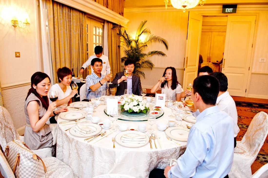 People sitting around a round dining table in a formal setting, each holding a glass, likely celebrating with drinks, in a luxurious room with elegant decor and a large floral centerpiece.