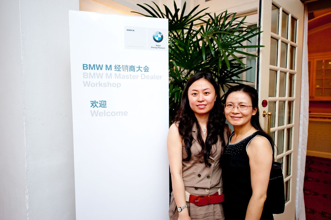 Two women standing next to a BMW Master Dealer Workshop sign at an event, with one woman wearing a beige outfit and the other wearing a black dress.
