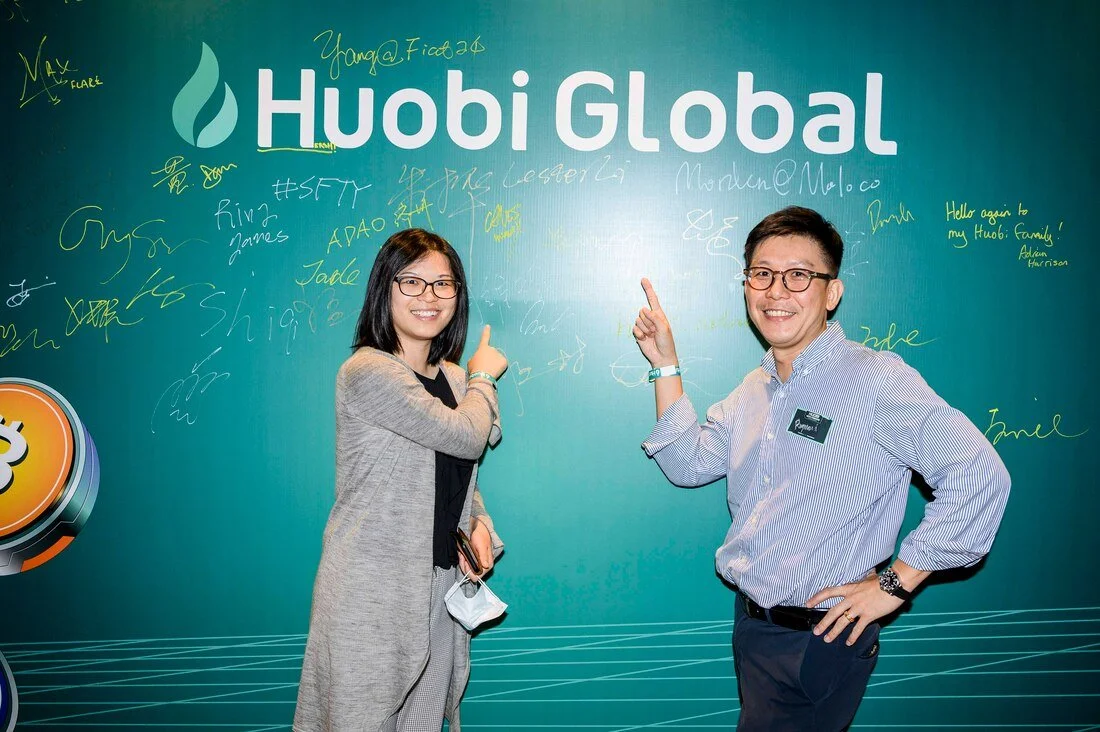 Two people, a woman and a man, smiling and pointing at a wall with the Huobi Global logo and multiple signatures or messages written on it. The woman has shoulder-length dark hair and glasses, wearing a gray cardigan. The man has short dark hair, gla