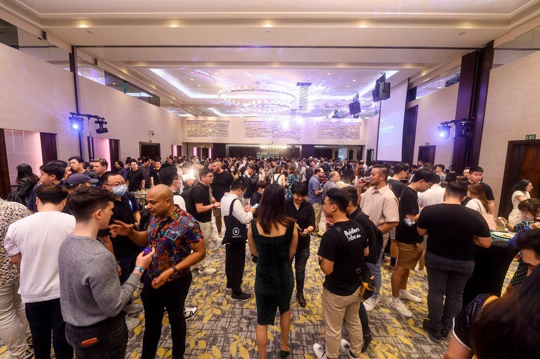 Crowd of people at a conference or event in a large, well-lit hall with screens and seating area.