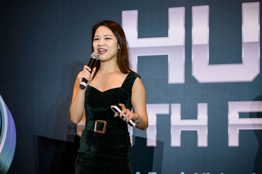 Woman speaking into a microphone on stage with a dark background and large white text behind her.
