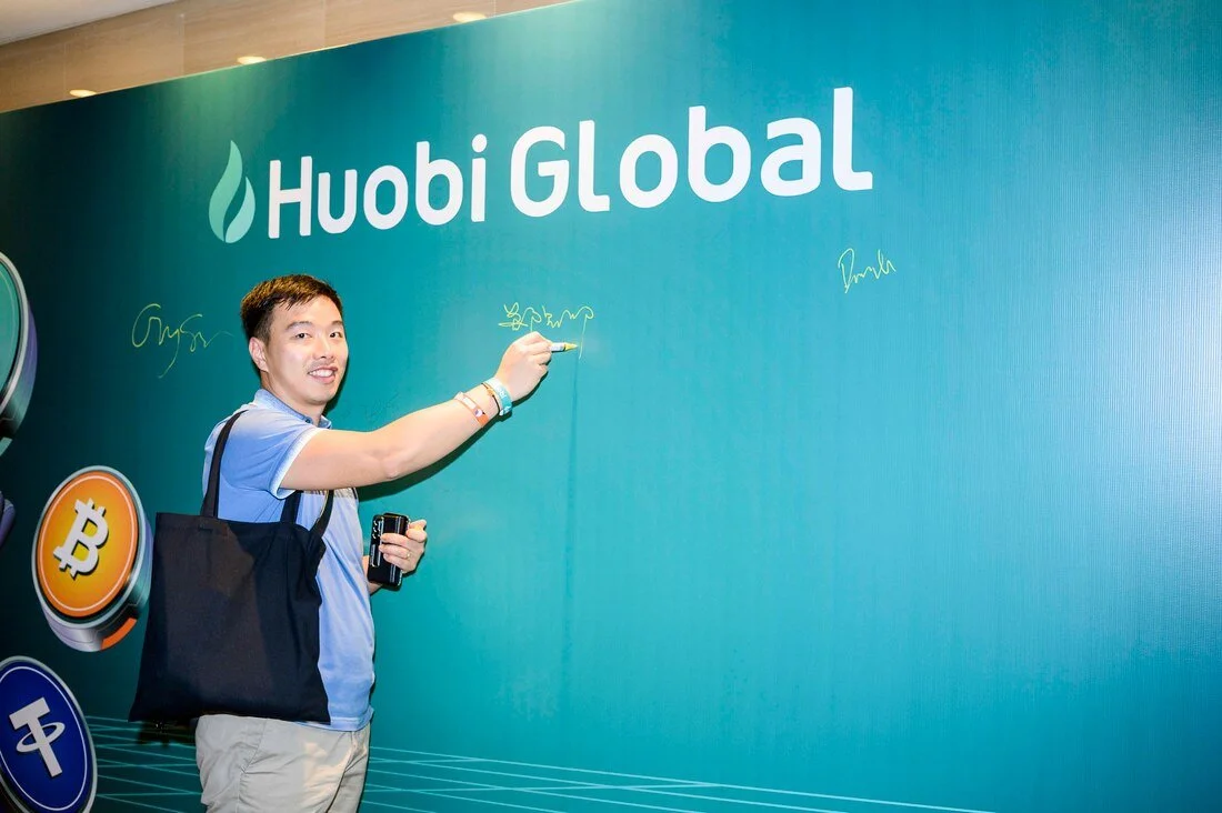 A man signing a large poster that reads "Huobi Global" with cryptocurrency symbols on the side