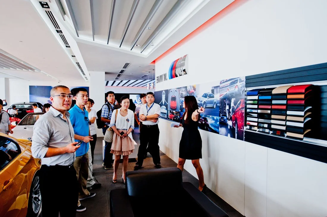 A group of people listening to a woman giving a presentation in a BMW showroom with car images and color samples on the wall.