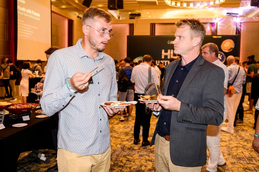Two men are conversing at a buffet-style event, holding plates of food, with a large crowd in the background.