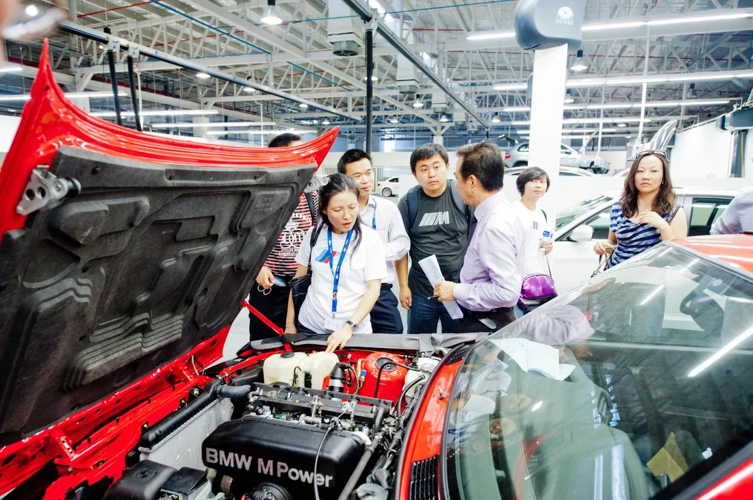 Group of people inspecting the engine of a red BMW sports car at an indoor auto exhibition.