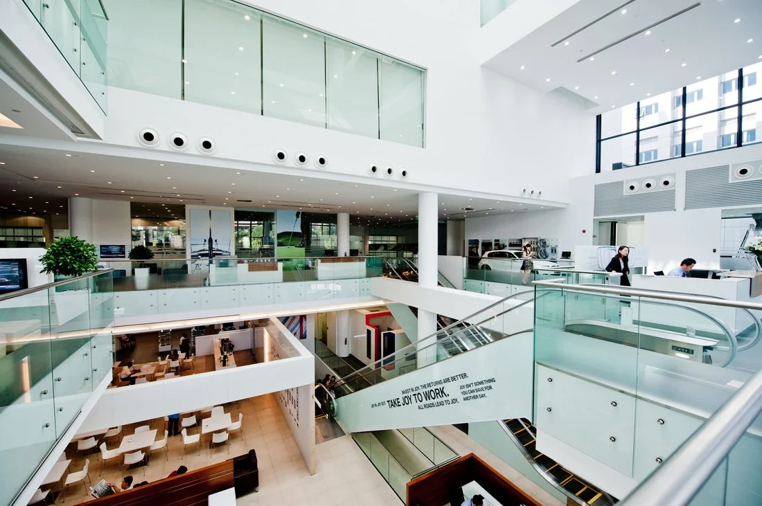 Interior of a modern, multi-level shopping mall or commercial building with glass railings, escalators, and large windows allowing natural light.