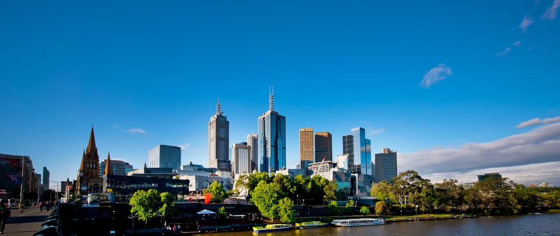 Skyline of Melbourne, Australia, with modern skyscrapers and a church, alongside the Yarra River, under a clear blue sky.