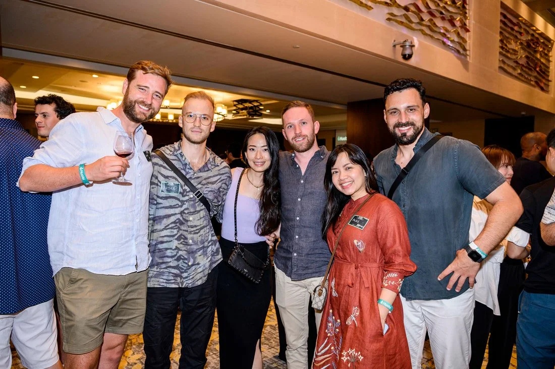 Group of six people smiling at a social event, some holding drinks, indoors with warm lighting and other attendees in the background.
