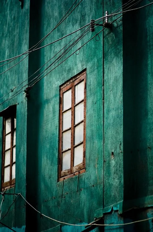 Close-up of an old teal-colored building with weathered wooden window frames and multiple hanging and running electrical wires.