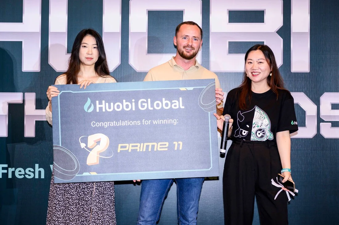 Three people on stage holding a large Huobi Global check for winning Prime 11; one woman holding a microphone, a man in the middle, and a woman on the right in black clothing.