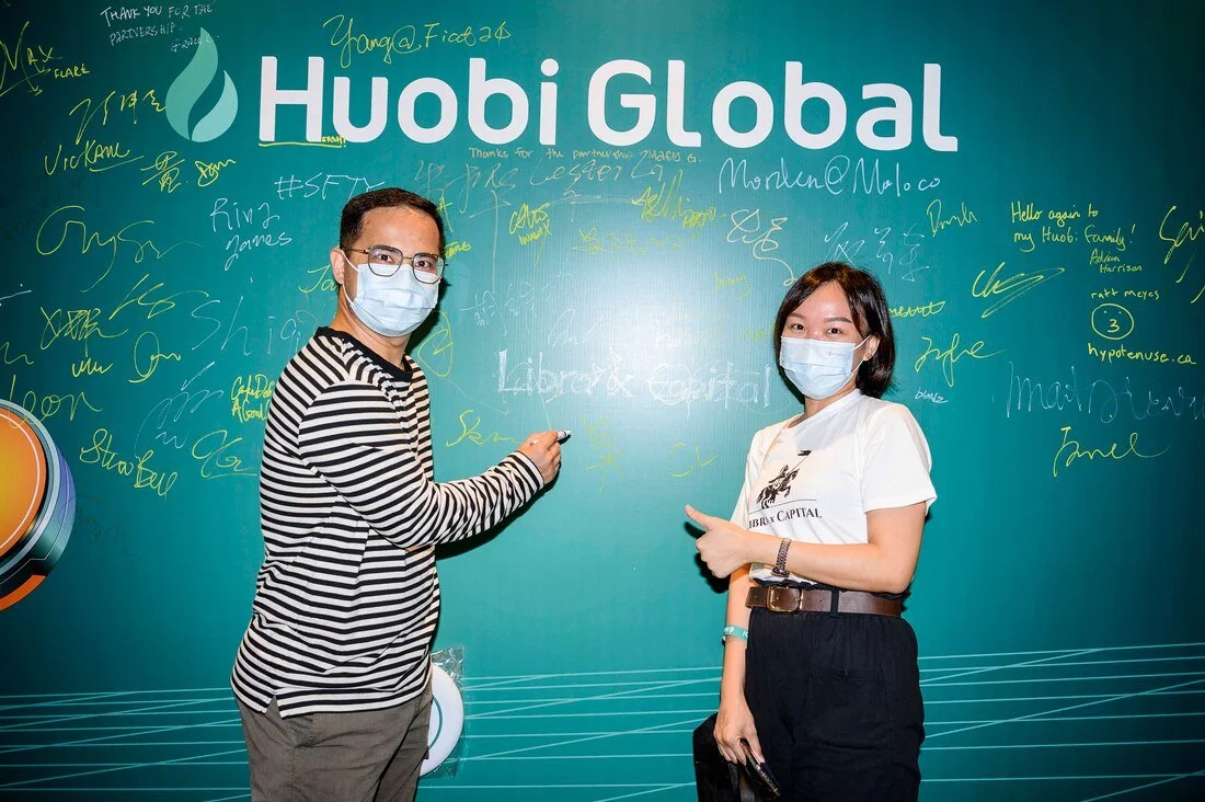 Two people wearing masks standing in front of a blue wall with signatures and notes, with the logo "Huobi Global" at the top, one man signing the wall and a woman pointing at it.